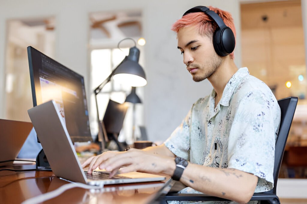 Asian man wearing headphones working on laptop at startup office. Young IT professional working at coworking office. Headphones vs earbuds