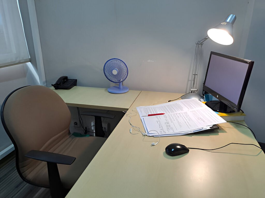 A well-lit, compact home office lighting featuring a wooden desk, ergonomic chair, computer monitor, and a modern desk lamp providing illumination. The setup includes a mouse, papers, and a small personal fan, suggesting a functional and comfortable area for work or study. Ideal for themes related to remote work, small businesses, productivity, or home office design